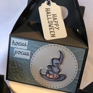 May include: Black Halloween gift box with a tag saying "HAPPY HALLOWEEN." The box has a witch's hat graphic and the words "hocus pocus." The box has a handle and is on a white surface.