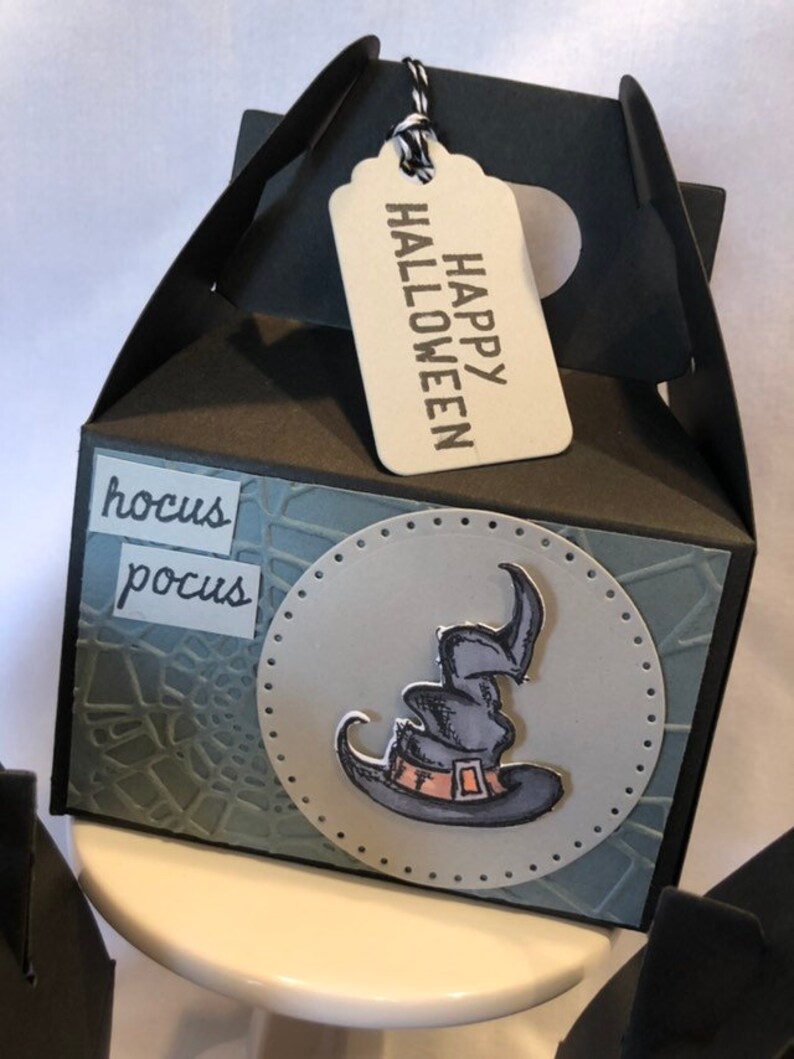 May include: Black Halloween gift box with a tag saying "HAPPY HALLOWEEN." The box has a witch's hat graphic and the words "hocus pocus." The box has a handle and is on a white surface.