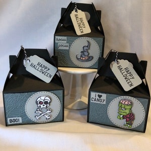 May include: Three black Halloween gift boxes, each with a "Happy Halloween" tag. The boxes display a skull and crossbones, a witch's hat, and Frankenstein's monster. The boxes also feature the words "Boo!" and "I ❤️ Candy."
