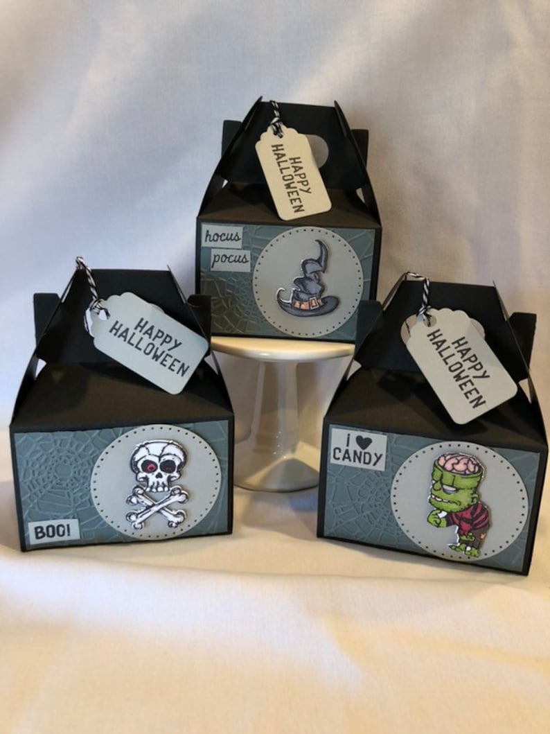 May include: Three black Halloween gift boxes, each with a "Happy Halloween" tag. The boxes display a skull and crossbones, a witch's hat, and Frankenstein's monster. The boxes also feature the words "Boo!" and "I ❤️ Candy."