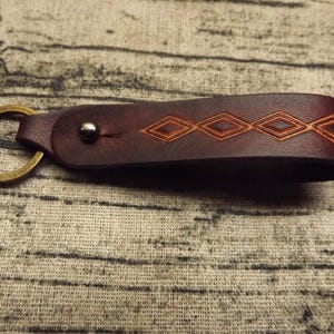 May include: A dark brown leather keyring with a brass ring and a diamond pattern. The leather is stitched and riveted, showcasing a handcrafted design. The keyring is a practical accessory.
