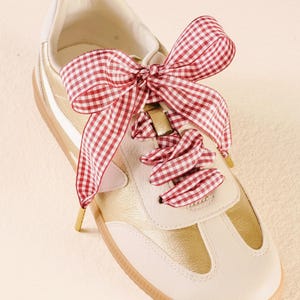 May include: A gold and white sneaker with a red and white gingham ribbon bow. The shoe has a white toe cap and a gold metallic finish on the sides. The laces are replaced with a decorative bow.