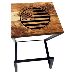 May include: A wooden bar stool with a black metal frame. The top of the stool is made of wood and has a circular design with the words "United States of America" and "Est. 1776" engraved in it. The design also includes a stylized American flag.
