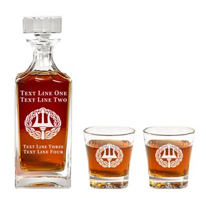 May include: A clear glass decanter with a square base and a stopper. The decanter is filled with amber liquid and has a white design of a trident and laurel wreath. The text "TEXT LINE ONE TEXT LINE TWO" is engraved on the front of the decanter. The text "TEXT LINE THREE TEXT LINE FOUR" is engraved on the bottom of the decanter. Two clear glass tumblers with square bases are also shown. The tumblers are filled with amber liquid and have a white design of a trident and laurel wreath.