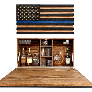 May include: A wooden cabinet with a hidden compartment that opens to reveal a bar area. The cabinet is decorated with a thin blue line American flag. The bar area is stocked with bottles of liquor, glasses, and a decanter.