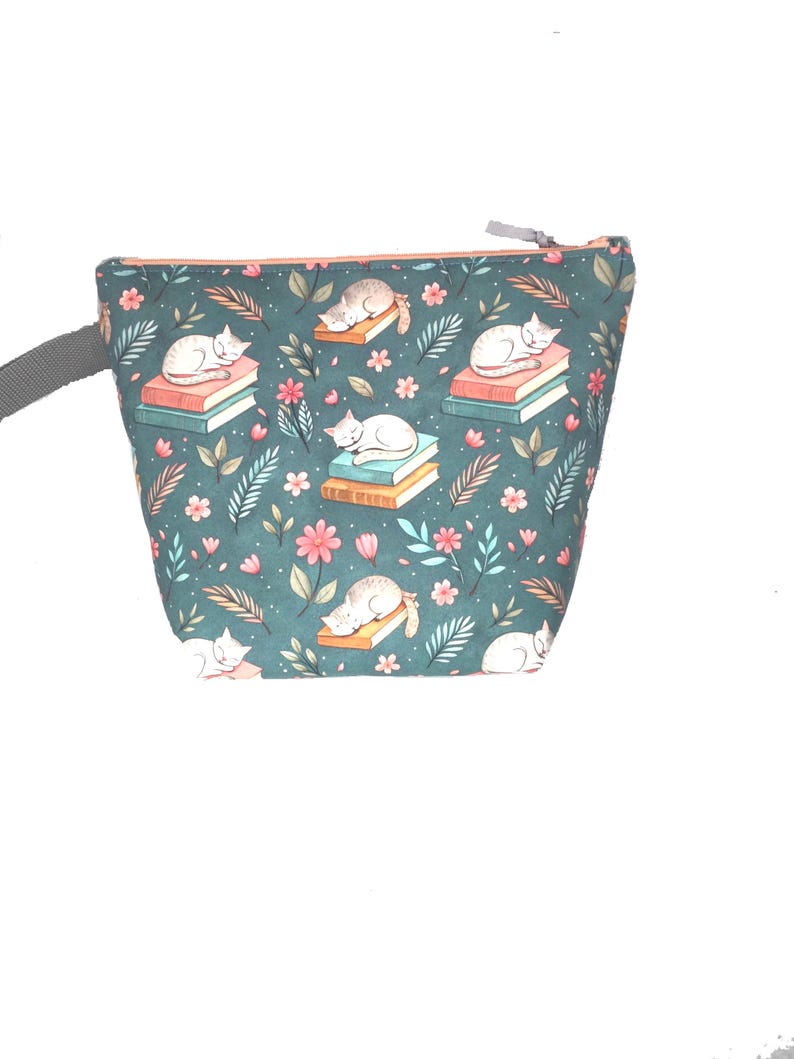 May include: A teal zippered pouch with a repeating pattern of sleeping white and gray cats on stacks of books, surrounded by pink flowers and green foliage. The pouch has a gray strap and an orange zipper.