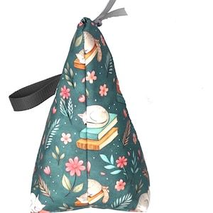May include: A teal fabric doorstop with a repeating pattern of sleeping white cats, books, and pink flowers. The doorstop has a gray handle and a gray ribbon at the top. The doorstop is cone-shaped.