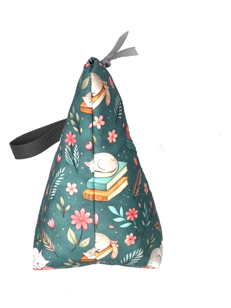 May include: A teal fabric doorstop with a repeating pattern of sleeping white cats, books, and pink flowers. The doorstop has a gray handle and a gray ribbon at the top. The doorstop is cone-shaped.