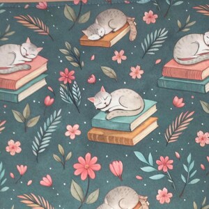 May include: A teal fabric with a repeating pattern of sleeping gray cats on stacks of books, surrounded by pink flowers and green and orange leaves. The design has a whimsical, storybook feel.