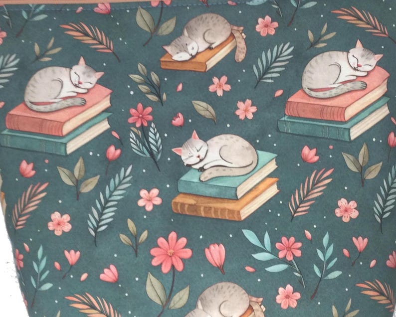 May include: A teal fabric with a repeating pattern of sleeping gray cats on stacks of books, surrounded by pink flowers and green and orange leaves. The design has a whimsical, storybook feel.
