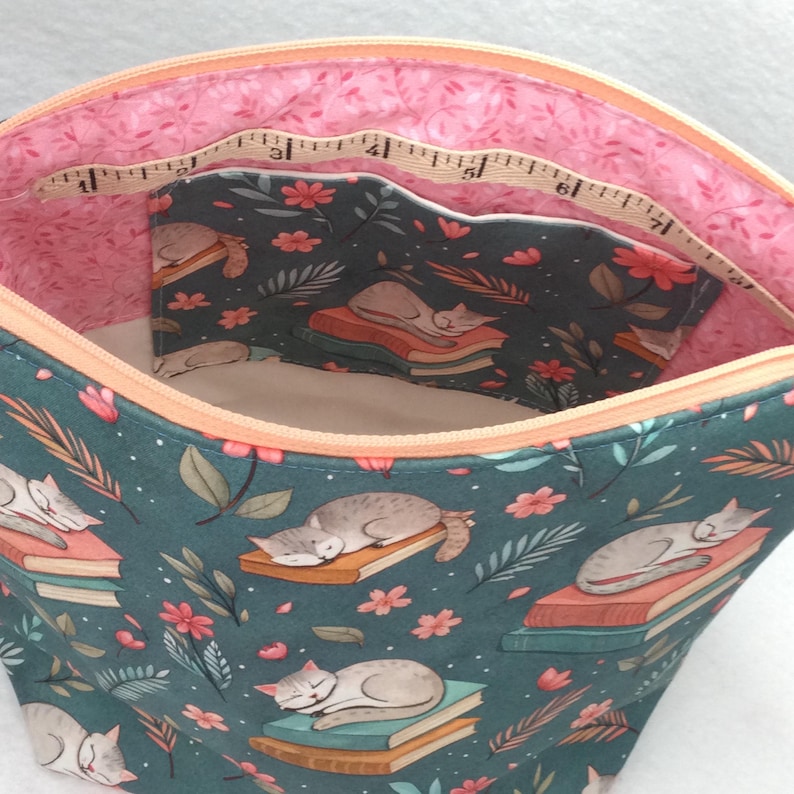 May include: A teal fabric pouch with a cat and book pattern. The interior features a pink floral lining and a small pocket with the same cat and book design. A measuring tape is visible inside the pouch.