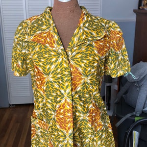 Puede incluir: Una camisa de manga corta con un llamativo estampado floral en amarillo, verde, blanco y marrón. La camisa se exhibe en un maniquí, destacando su ajuste a medida y diseño. Cuenta con cuello y dos bolsillos delanteros.