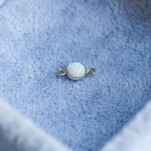 Puede incluir: Un peque&ntilde;o componente de joyer&iacute;a dorado con una piedra de &oacute;palo blanca y redonda en el centro. El &oacute;palo tiene una calidad sutil e iridiscente. El componente tiene peque&ntilde;os bucles a cada lado. El art&iacute;culo est&aacute; sobre un fondo azul claro y suave.