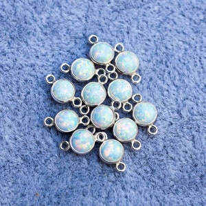 Puede incluir: Un conjunto de componentes de joyer&iacute;a redondos, plateados, con cabujones iridiscentes y opalinos. Cada componente tiene un peque&ntilde;o lazo para la fijaci&oacute;n. Los componentes est&aacute;n dispersos sobre una superficie azul texturizada.