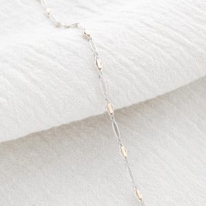 May include: A delicate silver necklace with alternating oval and diamond-shaped links, accented with small gold-colored details. The necklace is displayed on a white, textured fabric, creating a soft, elegant aesthetic. The chain is thin and appears lightweight.