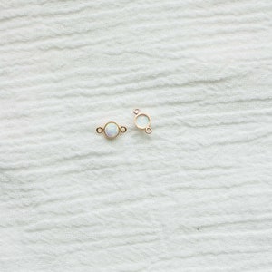 Puede incluir: Dos conectores de joyer&iacute;a dorados con una piedra redonda iridiscente similar a un &oacute;palo en el centro. Los conectores tienen peque&ntilde;os bucles a cada lado para la fijaci&oacute;n. El fondo es una tela blanca texturizada.