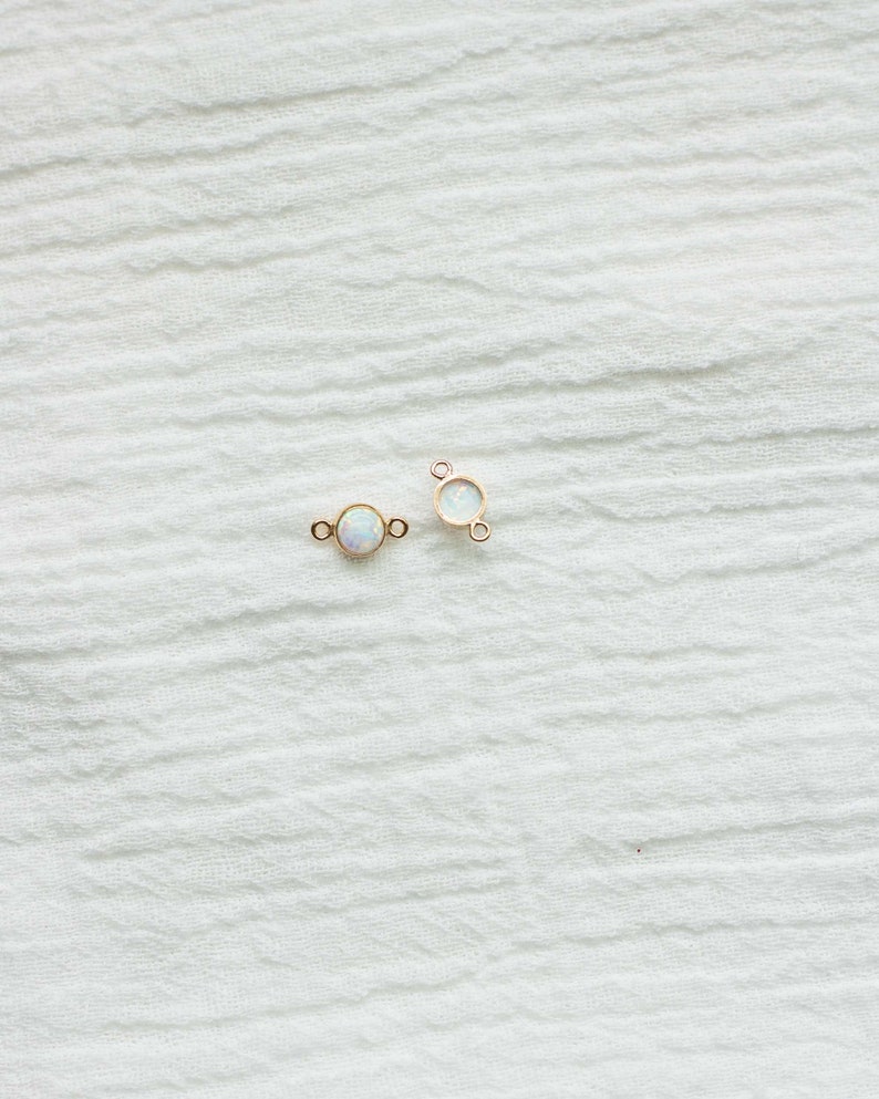 Puede incluir: Dos conectores de joyer&iacute;a dorados con una piedra redonda iridiscente similar a un &oacute;palo en el centro. Los conectores tienen peque&ntilde;os bucles a cada lado para la fijaci&oacute;n. El fondo es una tela blanca texturizada.