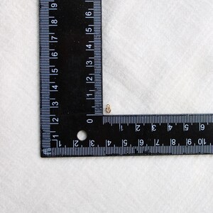 Puede incluir: Un peque&ntilde;o dije en forma de coraz&oacute;n dorado se encuentra junto a una regla negra. La regla muestra marcas blancas para cent&iacute;metros. El dije mide aproximadamente 0,6 cm. El fondo es una tela blanca apagada.