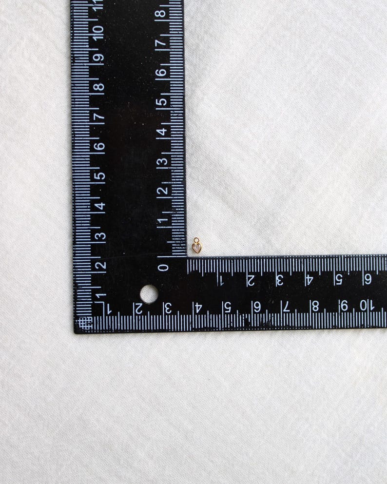 Puede incluir: Un peque&ntilde;o dije en forma de coraz&oacute;n dorado se encuentra junto a una regla negra. La regla muestra marcas blancas para cent&iacute;metros. El dije mide aproximadamente 0,6 cm. El fondo es una tela blanca apagada.