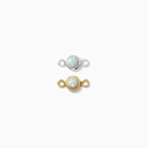 May include: Two small opal connectors, one silver and one gold, are displayed against a white background. Each connector features a round opal stone set in a bezel setting with small loops on either side for attachment. The gold connector has a warm, yellow hue.