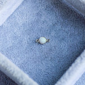 Puede incluir: Un peque&ntilde;o dije redondo de &oacute;palo con un engaste dorado. El &oacute;palo tiene un aspecto blanco lechoso. El dije se muestra sobre una superficie azul claro texturizada, posiblemente terciopelo o fieltro. El dije tiene dos peque&ntilde;os bucles a cada lado.