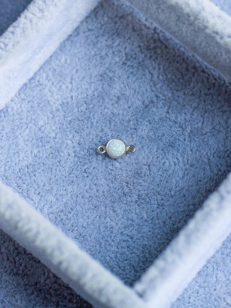 Puede incluir: Un peque&ntilde;o dije redondo de &oacute;palo con un engaste dorado. El &oacute;palo tiene un aspecto blanco lechoso. El dije se muestra sobre una superficie azul claro texturizada, posiblemente terciopelo o fieltro. El dije tiene dos peque&ntilde;os bucles a cada lado.