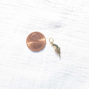 Pode incluir: Um amuleto dourado em forma de pistola, ao lado de um centavo americano para compara&ccedil;&atilde;o de tamanho.