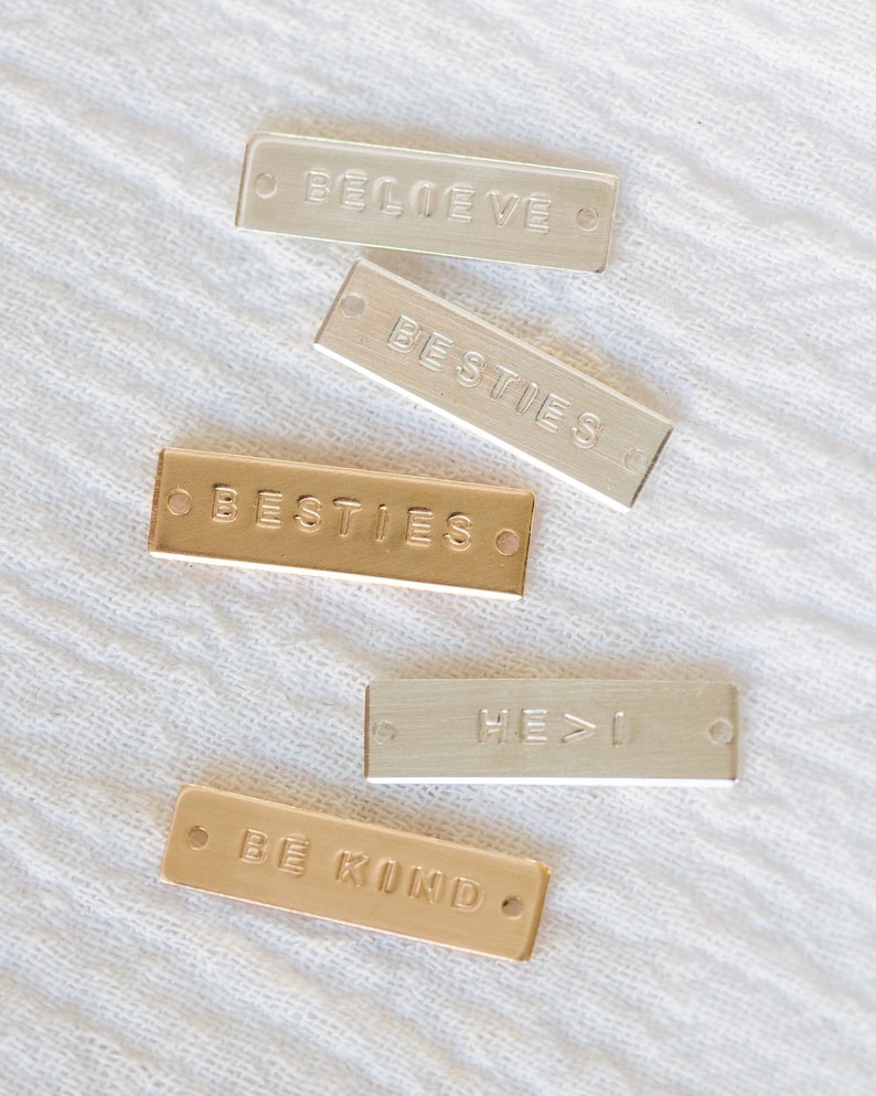 K&ouml;nnte beinhalten: F&uuml;nf rechteckige Metallanh&auml;nger mit gepr&auml;gtem Text. Die Anh&auml;nger sind silber- und goldfarben und tragen die Worte "BELIEVE", "BESTIES", "BESTIES", "HE > I" und "BE KIND". Jeder Anh&auml;nger hat an jedem Ende ein kleines Loch.