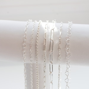 May include: Six silver chains of different styles and lengths displayed on a white surface.