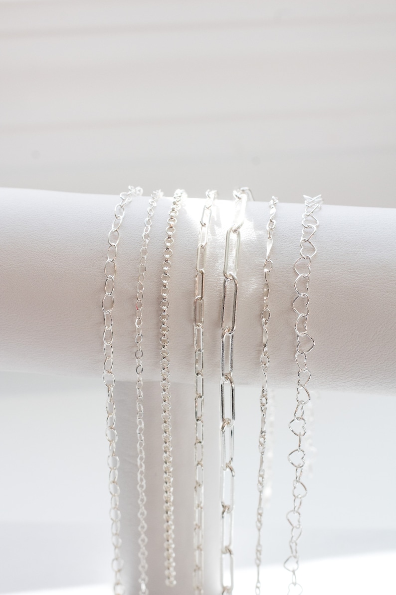 May include: Six silver chains of different styles and lengths displayed on a white surface.