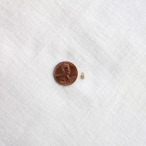 Puede incluir: Un peque&ntilde;o dije en forma de coraz&oacute;n dorado con una peque&ntilde;a piedra transparente, junto a una moneda de un centavo para comparar el tama&ntilde;o. El dije est&aacute; sobre una superficie blanca texturizada.