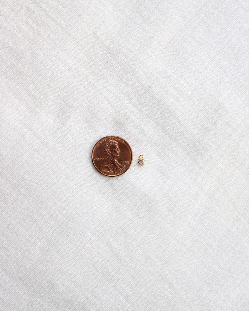 Puede incluir: Un peque&ntilde;o dije en forma de coraz&oacute;n dorado con una peque&ntilde;a piedra transparente, junto a una moneda de un centavo para comparar el tama&ntilde;o. El dije est&aacute; sobre una superficie blanca texturizada.