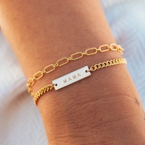K&ouml;nnte beinhalten: Zwei goldfarbene Armb&auml;nder sind abgebildet. Eines hat ein Kettenglied-Design, das andere eine rechteckige Platte mit dem eingravierten Wort "MAMA". Die Armb&auml;nder befinden sich an einem Handgelenk, der Hintergrund ist unscharf.