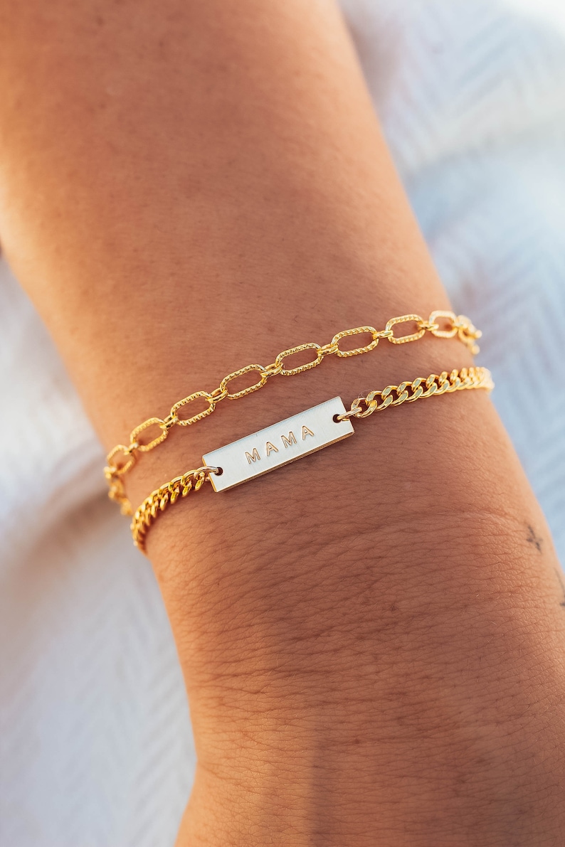 K&ouml;nnte beinhalten: Zwei goldfarbene Armb&auml;nder sind abgebildet. Eines hat ein Kettenglied-Design, das andere eine rechteckige Platte mit dem eingravierten Wort "MAMA". Die Armb&auml;nder befinden sich an einem Handgelenk, der Hintergrund ist unscharf.