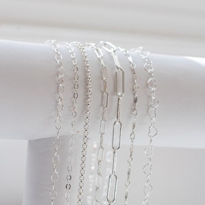 May include: A collection of six silver chains of different styles and lengths, displayed on a white surface.
