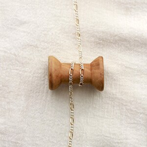 May include: A silver Figaro chain necklace hangs from a wooden spool. The chain features a repeating pattern of elongated and circular links. The spool is made of light brown wood and is set against a white fabric background.