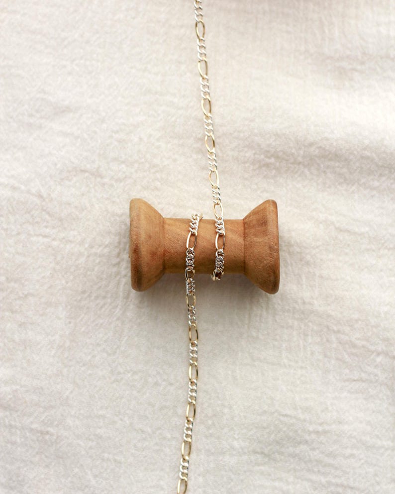 May include: A silver Figaro chain necklace hangs from a wooden spool. The chain features a repeating pattern of elongated and circular links. The spool is made of light brown wood and is set against a white fabric background.