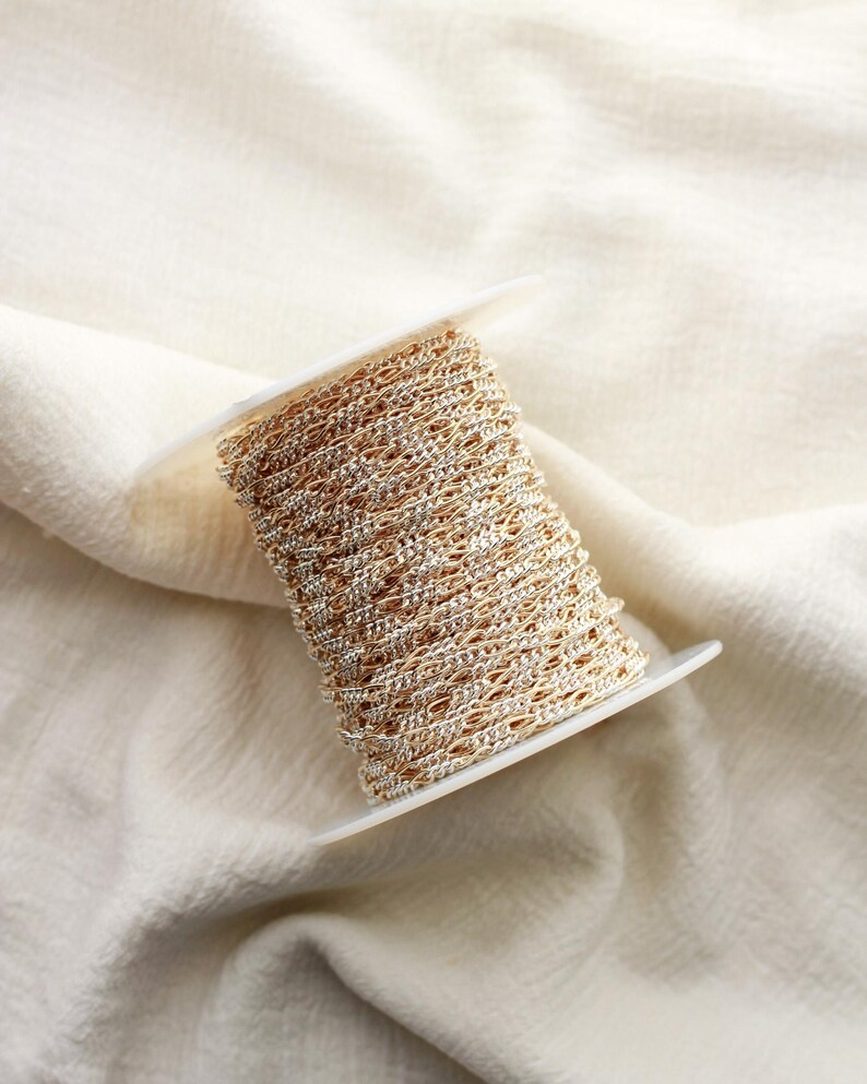 May include: A spool of gold-coloured chain with silver detailing. The chain is wound on a white plastic spool. The chain is suitable for jewellery making or craft projects.
