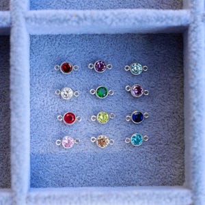 May include: A close-up view of a jewelry box with 12 small silver charms, each with a different colored gemstone. The gemstones are red, purple, blue, green, white, and yellow.