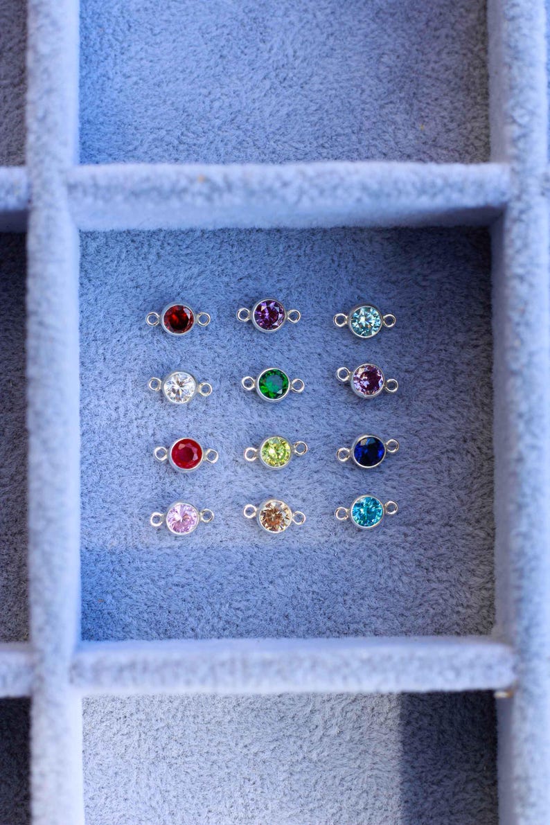 May include: A close-up view of a jewelry box with 12 small silver charms, each with a different colored gemstone. The gemstones are red, purple, blue, green, white, and yellow.