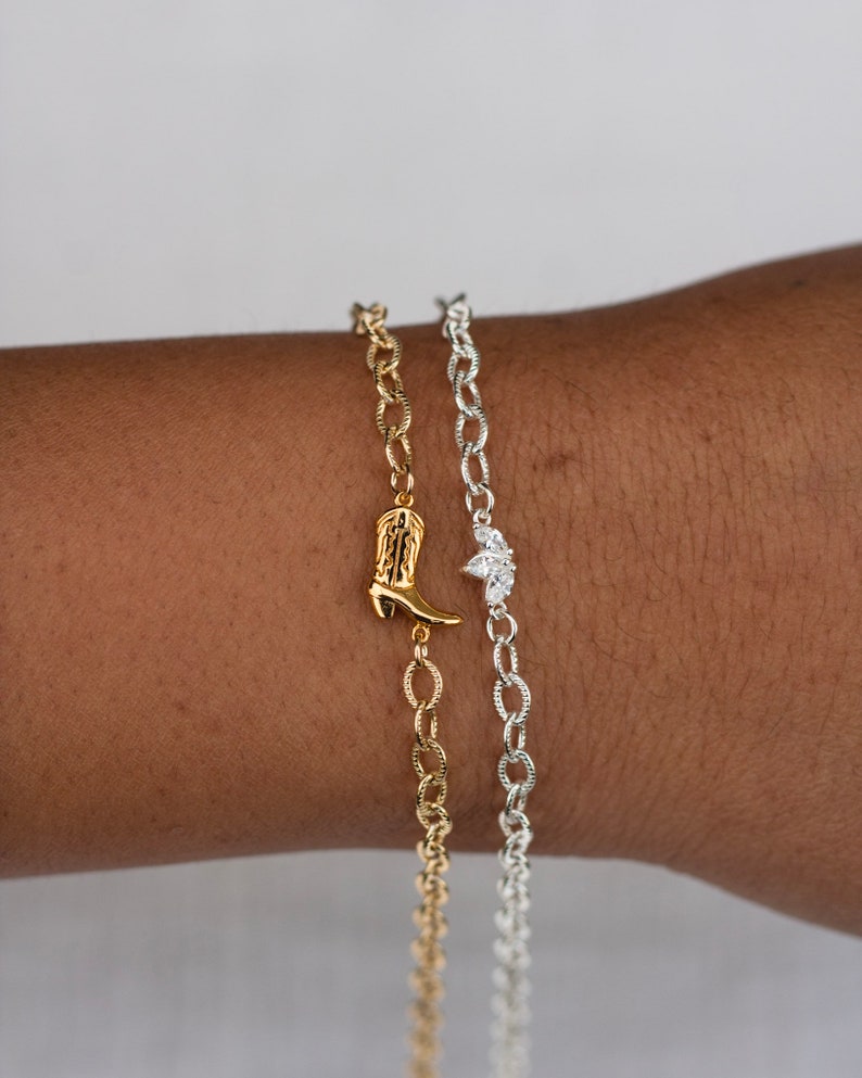 Op de afbeelding: Twee gouden kettingarmbanden met bedels. De ene armband heeft een kleine laarsbedel, de andere heeft een bedel met drie kleine diamanten.