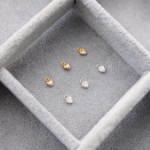 Puede incluir: Primer plano de peque&ntilde;os dijes en forma de coraz&oacute;n en tonos dorados y plateados, cada uno con un centro de piedra preciosa transparente. Los dijes est&aacute;n dispuestos dentro de una caja de joyer&iacute;a de terciopelo gris, mostrando su delicado dise&ntilde;o.