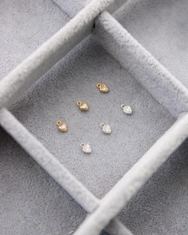 Puede incluir: Primer plano de peque&ntilde;os dijes en forma de coraz&oacute;n en tonos dorados y plateados, cada uno con un centro de piedra preciosa transparente. Los dijes est&aacute;n dispuestos dentro de una caja de joyer&iacute;a de terciopelo gris, mostrando su delicado dise&ntilde;o.