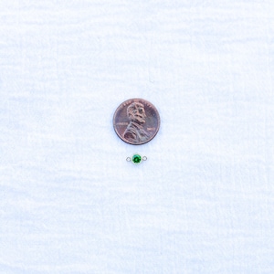 May include: A small green gemstone charm with silver jump rings. The charm is shown next to a US penny for size comparison.