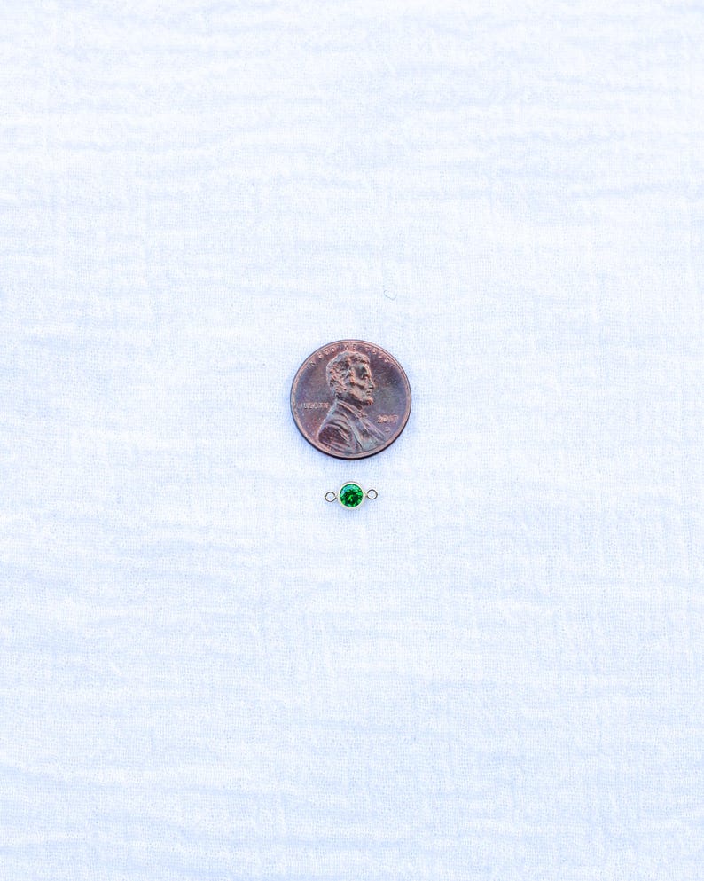 May include: A small green gemstone charm with silver jump rings. The charm is shown next to a US penny for size comparison.