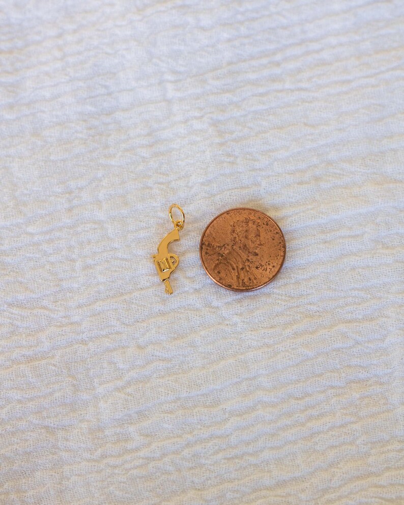 Pode incluir: Um amuleto dourado em forma de pistola, ao lado de um centavo americano.