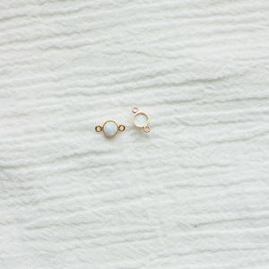 Puede incluir: Dos peque&ntilde;os conectores de joyer&iacute;a dorados con una piedra redonda iridiscente similar al &oacute;palo en el centro. Cada conector tiene un bucle a cada lado para sujetar a otros componentes de joyer&iacute;a. Se muestran sobre una tela blanca texturizada.