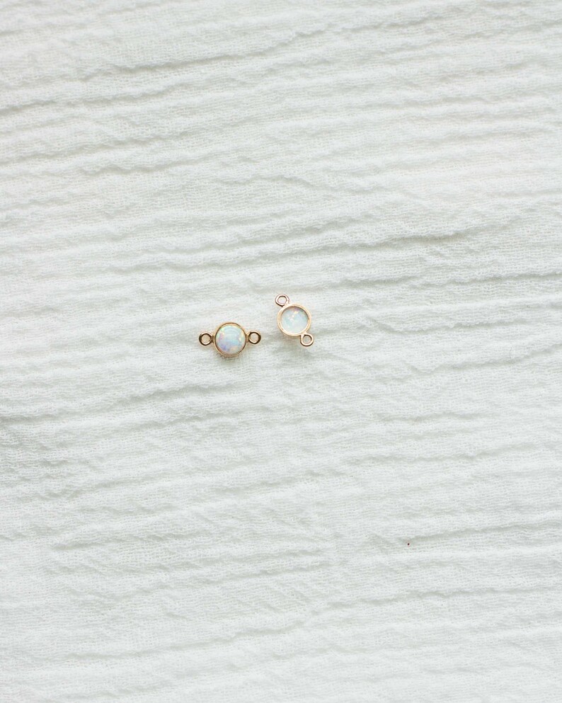 Puede incluir: Dos peque&ntilde;os conectores de joyer&iacute;a dorados con una piedra redonda iridiscente similar al &oacute;palo en el centro. Cada conector tiene un bucle a cada lado para sujetar a otros componentes de joyer&iacute;a. Se muestran sobre una tela blanca texturizada.