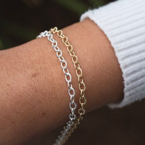 Op de afbeelding: Twee kettingarmbanden, een zilveren en een gouden, om een pols. Beide armbanden zijn gemaakt van een delicate ketting met ovale schakels.