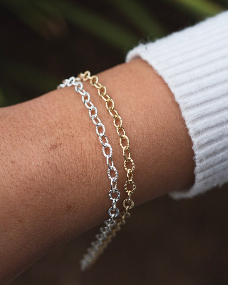 Op de afbeelding: Twee kettingarmbanden, een zilveren en een gouden, om een pols. Beide armbanden zijn gemaakt van een delicate ketting met ovale schakels.